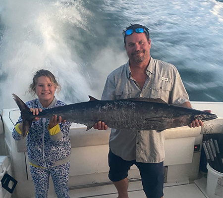 A man and a young girl stand next to one another while they both hold a Wahoo (a large fish) while standing on a boat over the water.