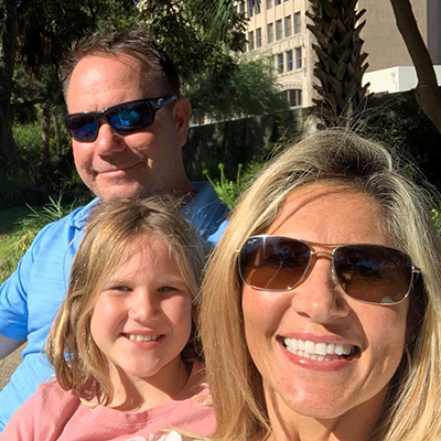 A man, woman, and young girl all sit closely to one another as the man and woman wear sunglasses and they all smile at the camera.