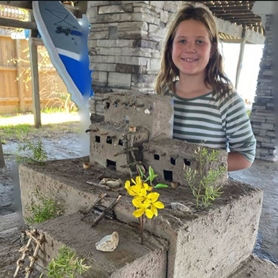 A young girl stands outdoors behind a handmade structure with multiple levels, flowers, and grass.