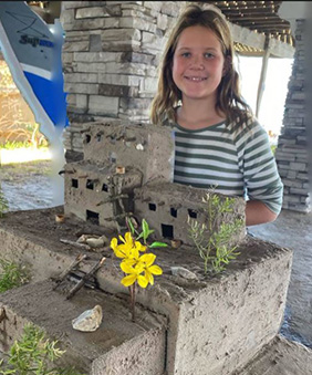 A young girl stands outdoors behind a handmade structure with multiple levels, flowers, and grass.