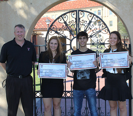 A man stands next to a young man and two young women outdoors as the young people all hold large checks in their hands.