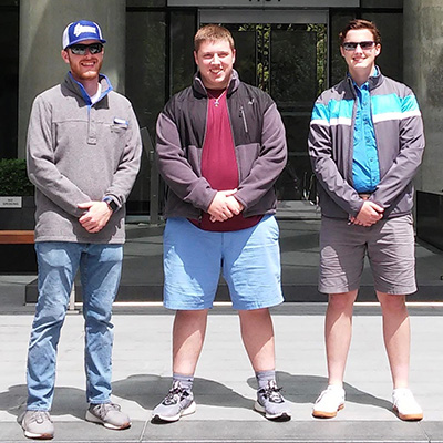 Three young men stand side by side outside wearing jackets and smiling at the camera.