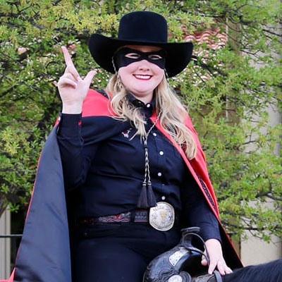A woman sits on the back of a black horse while wearing a black mask, red and black cape and cowboy boots.