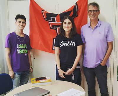 A young woman stands between a younger man to her right and an older man to her left as all three stand indoors between a desk and a Texas Tech flag.