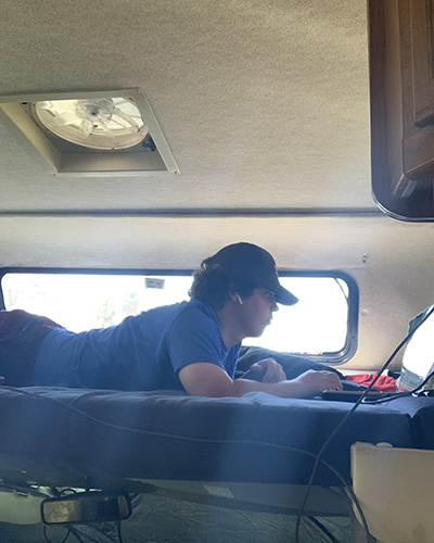 A teenage boy lies on his stomach on a bed while wearing a hat and looking at his laptop screen.