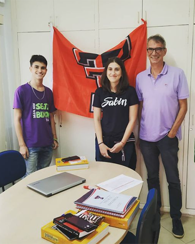 A young woman stands between a younger man to her right and an older man to her left as all three stand indoors between a desk and a Texas Tech flag.