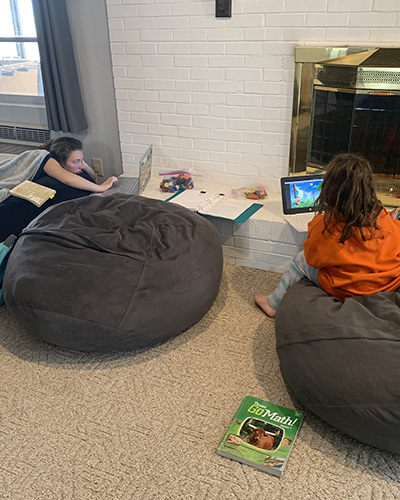 Serena and Kaydence do schoolwork at an Air BnB. A teenage girl lies on a carpeted floor with one hand on her keyboard while staring at a computer screen while another, younger, girl sits on a bean bag chair while watching a laptop screen.