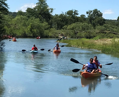 A group of students in various kayaks row down a river outdoors.