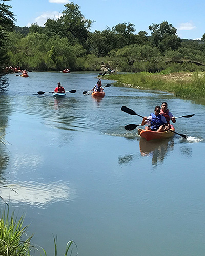 Numerous kayaks rest on a river with people sitting in them in an outdoor setting.