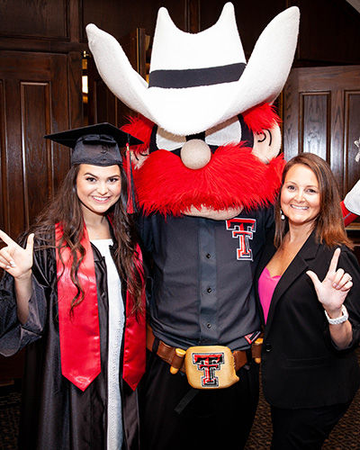 The Texas Tech mascot Raider Red stands between two women as all three perform the guns up sign.