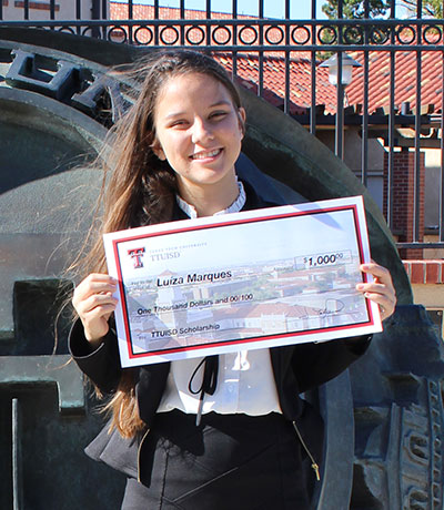 A young woman stands in front of a large structure of a Texas Tech ring while holding a large check in both hands.