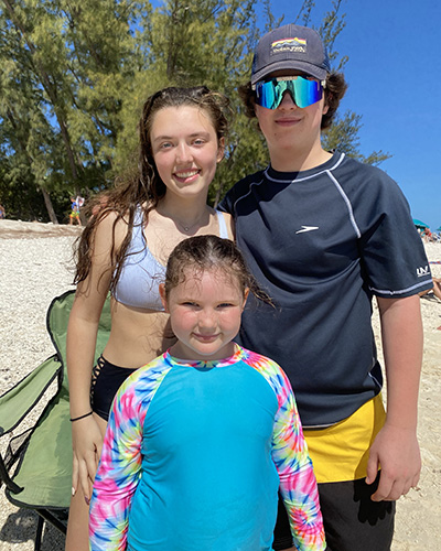 A teenage male and teenage female stand beside each other outdoors as a small child stands in front of them while they all smile wearing swim gear.