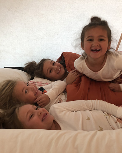 Four small, young girls lie beside one another on a large pillow under a covered tent with a large blanket covering three of the girls.