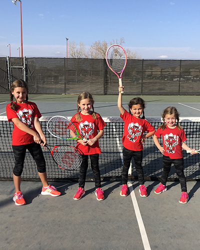 Four small, young girls stand next to one another all wearing the same texas tech shirts, black bottoms, and red shoes outdoors while holding tennis rackets.