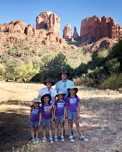 Four small, young girls stand side-by-side as a woman and man stand behind them with the background of trees and a large canyon behind them all.