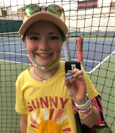 A young girl wearing a yellow cap and yellow shirt with a tennis racket on her back smiles and hold a small round pin in her left hand.