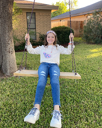 A young girl sits on a swing hanging from a front yard tree while smiling.