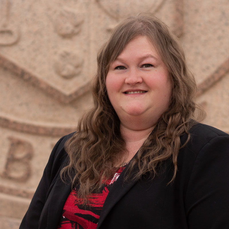 A woman wearing a black blazer and red shirt stands outdoors in front of the Texas Tech University seal.