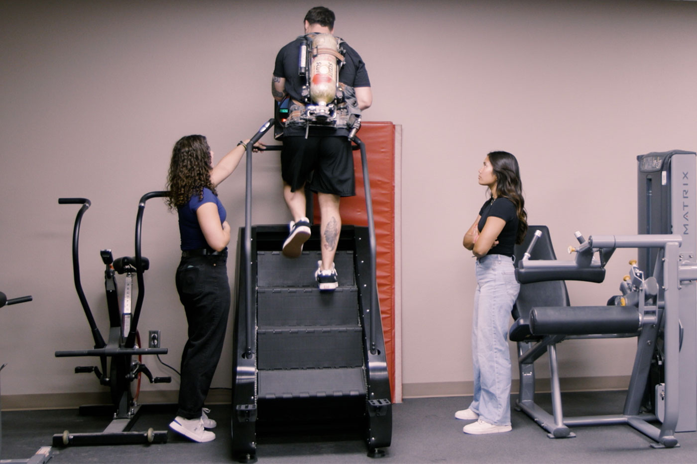 a student is on a stair master with an oxygen tank on his back while two other students monitor him