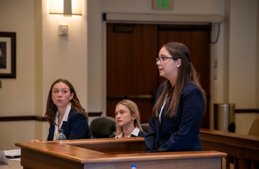 Kiera Figgins, a Texas Tech Law student, stands at the podium presenting oral argument during the Chapman, Lewis Clark Moot Court Invitational, with members of the opposing team visible in the background.