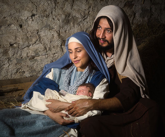 Live Christmas nativity scene in an old barn, actors portray Mary and Joseph holding a realistic doll representing baby Jesus.