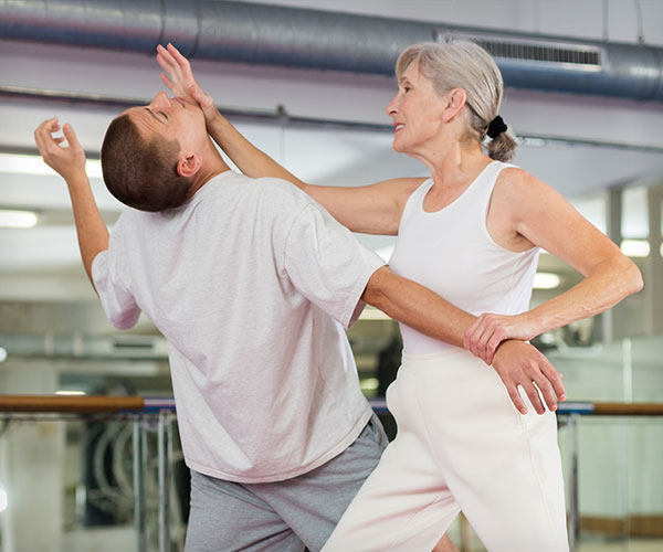 Older woman performing a palm heel strike to a young man's chin in a gym.