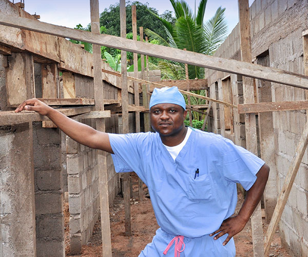 Sixtus Atabong in scrubs resting hand partially constructed wall of a hospital expansion in Muyuka.
