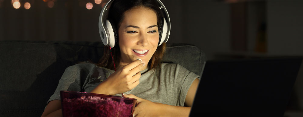  Smiling woman with headphones eating popcorn in front of a laptop in a dark room.