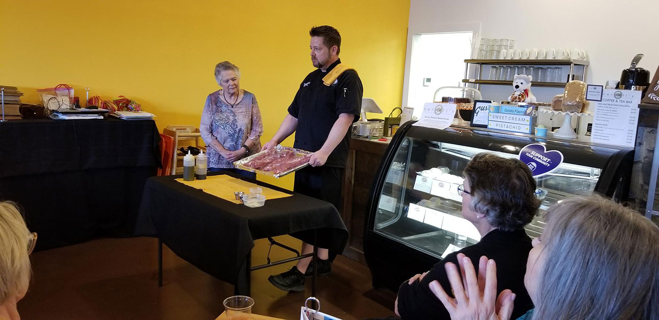 Chef Jason Beard prepares food to be cooked in front of OLLI members. Chef Beard stands behind a table with a black cloth drapped over it while holding a pan with slabs of meat on it and looks into the crowd of OLLI members as they watch him from the opposite side of the room.