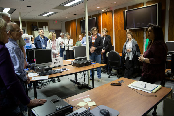 The LPD Call Center A group of OLLI members stand among computers and desks in a room and watch a speaker in the corner of the room