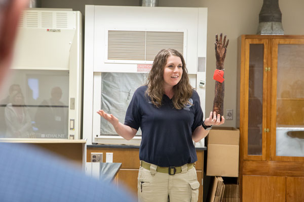 Evidence Replica A woman in a navy blue LPD shirt stands at the front of a room while holding a burned arm replica in her left hand with a red tag wrapped around it.