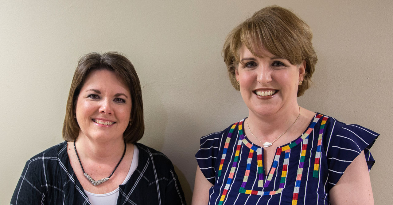 Tina Crowson (left) and Shelby Crews Tina and Shelby stand shoulder to shoulder and in front of a cream colored wall while smiling.