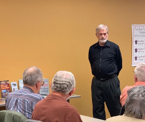 Ross Bigelow shares on how Fredericksburg impacts the world. Ross Bigelow stands at the front of a classroom facing his audience with his hands clasped together behind his back.