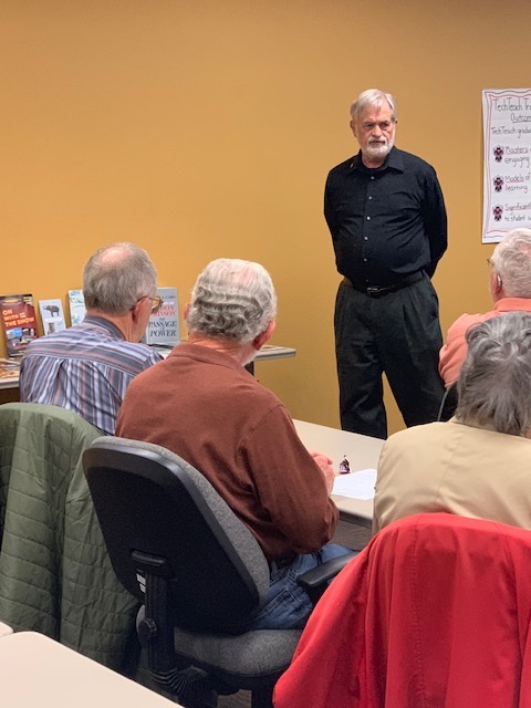 Ross Bigelow shares on how Fredericksburg impacts the world. Ross Bigelow stands at the front of a classroom facing his audience with his hands clasped together behind his back.