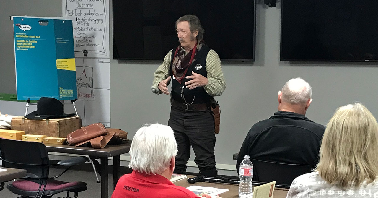 Former Texas Ranger Marvin Schroeder presents to OLLI members. Marvin Schroeder stands at the front of a classroom wearing a black vest and green and white stripped shirt while addressing his audience.