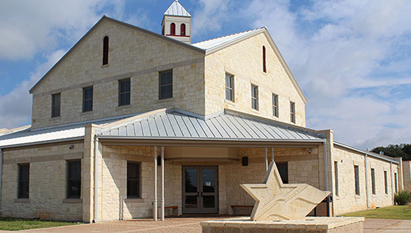 The Hill Country University Center in Fredericksburg, TX. A large brick building with numerous windows and a structure of a star at the front stand against a blue sky with clouds.