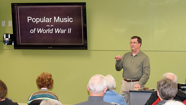 Larry Hess instructing a course to OLLI members Larry Hess stands at the front of a classroom, facing and motioning to his audience, with a mounted television screen behind him that reads"Popular Music of World War II".