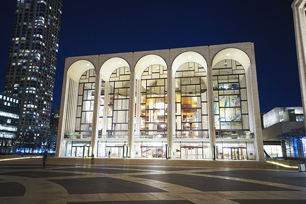 An white-colored opera house building is well-lit against a dark blue sky and stands near a tall, vertical building