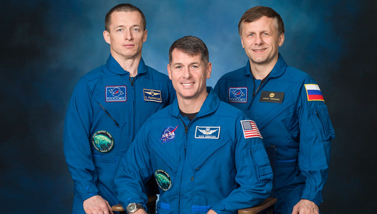 Kimbrough with crew members Andrei Borisenko and Oleg Novitskiy Kimbrough sits between two other men who are standing while all wear blue jumpsuits displaying their countries flag and logo for their space agency.