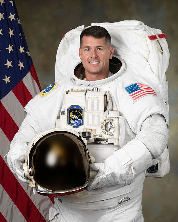 NASA astronaut Robert S. Kimbrough Kimbrough poses for a portrait while he stands next to an American flag wearing his spacesuit and holding his helmet and smiles at the camera