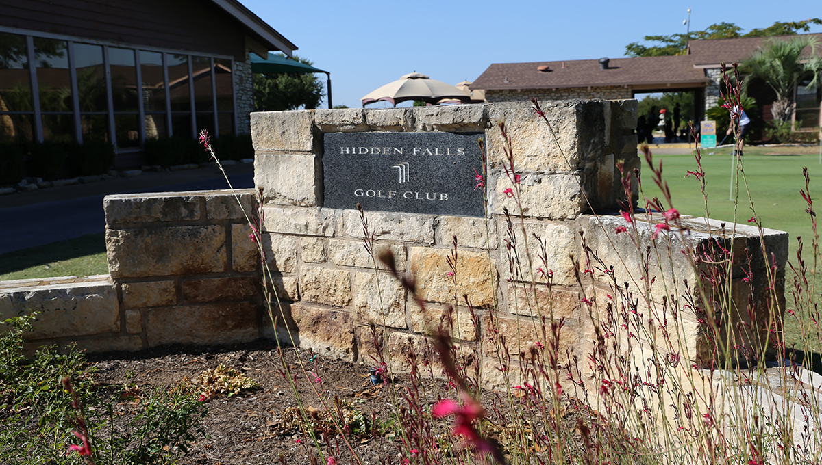 Hidden Falls Golf Club in Meadowlakes, TX A plaque that reads "Hidden Falls Golf Club" is encased in a stone fixture that is surrounded by areas of green grass, dirt and other plants with a clear blue sky in the background