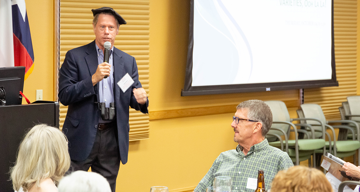 James Klein wears a black beret and binoculars James Klein stands at the front of a room wearing a black beret and holding a microphone to his mouth as his audience sits and look towards their speaker