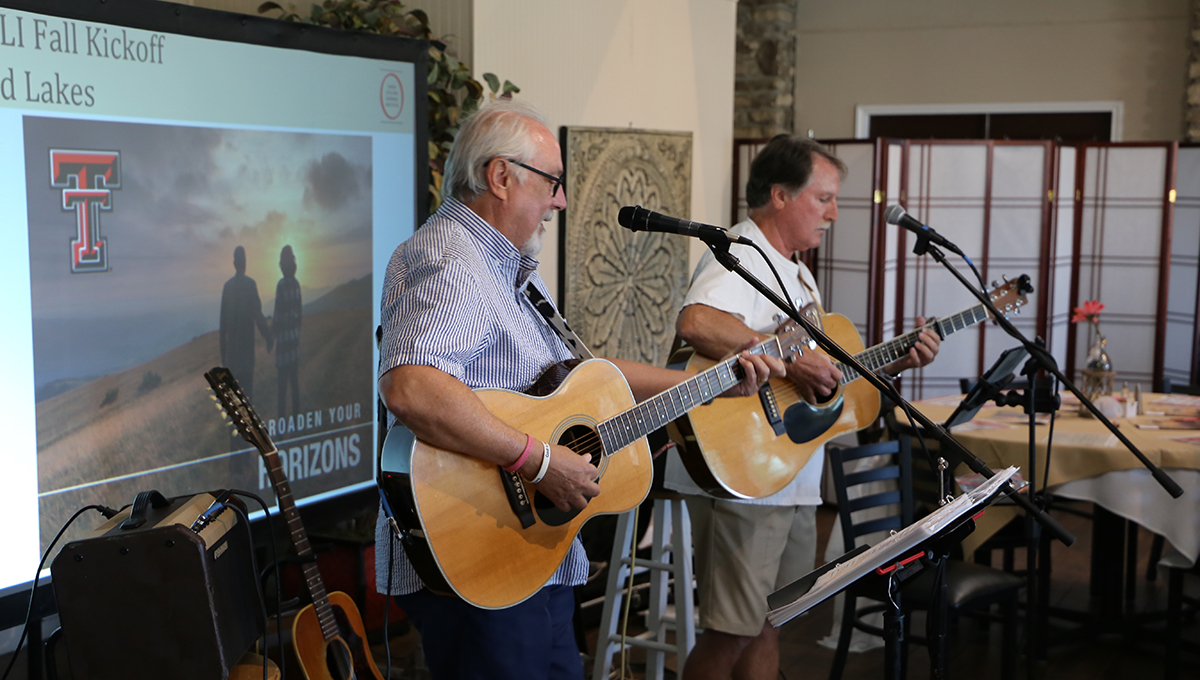 Highland Lakes kickoff entertainment Two men hold acoustic guitars in their hands and stand behind two microphones while looking down with a third guitar and a projector screen behind them