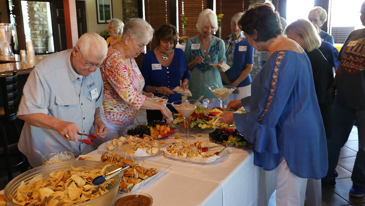 OLLI members enjoying the hors d'oeuvres buffet A group of men and women stand on both sides of a long table with white draping while leaning over and placing hors d'oeuvres on their plates