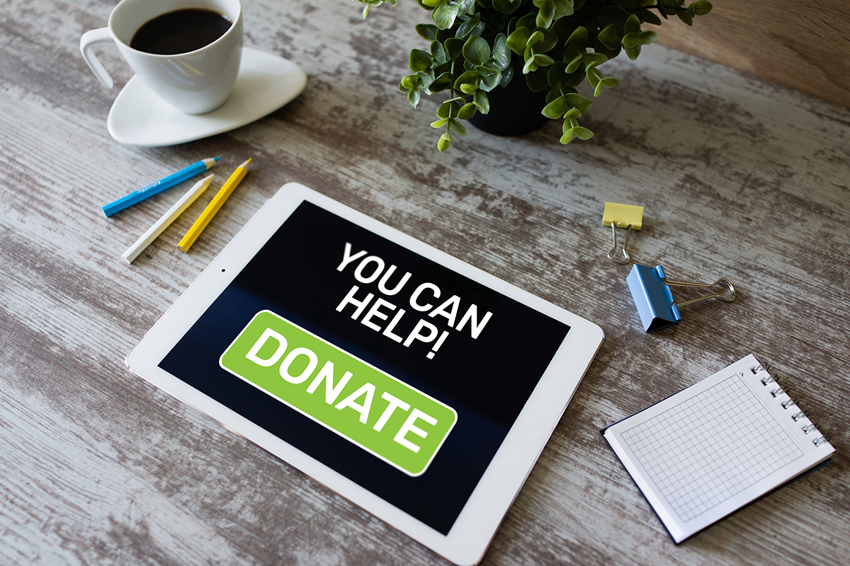 Donate A white tablet rests on a wooden table with the words "You Can Help! Donate" alongside a filled coffee cup, pencils, a plant, a notepad, and paper clips