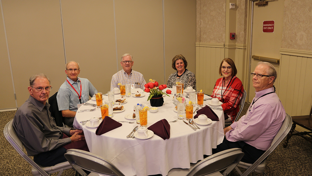 Older men and women sit around a round table with a white drape on top and they look at the camera and smile