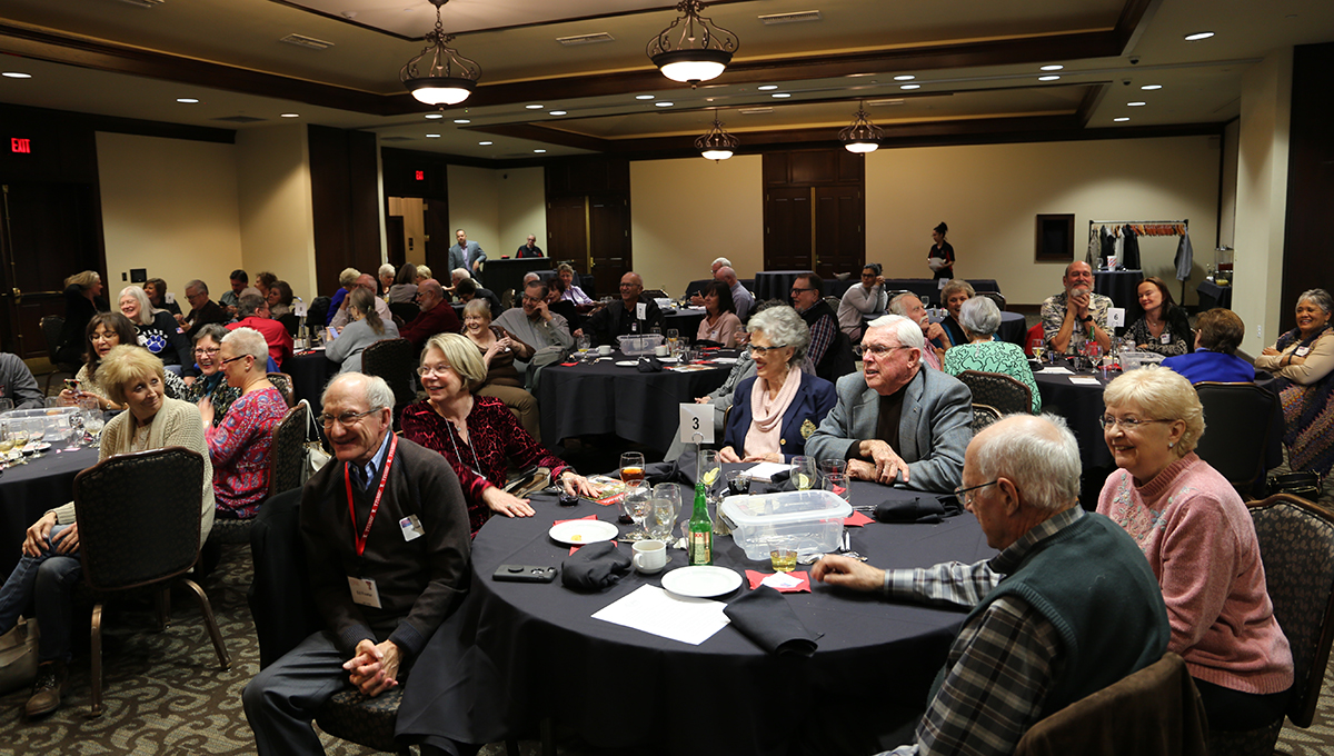 A large room is filled with numerous men and women sitting at round tables while all smile and look in different directions