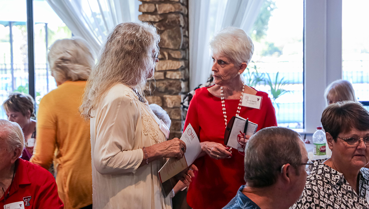 Elaine speaking with OLLI member at Highland Lakes Kickoff event Two older women stand next to one another facing each other in center frame among other people that are sitting