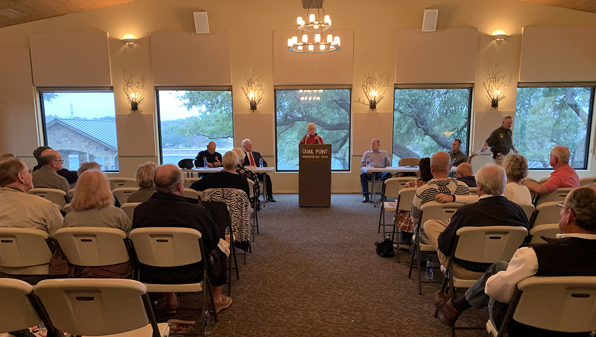 OLLI Vice Chair Elaine Waddill addressing the class A woman stands behind a podium and faces a large group of men and women as they sit in chairs inside an enclosed building