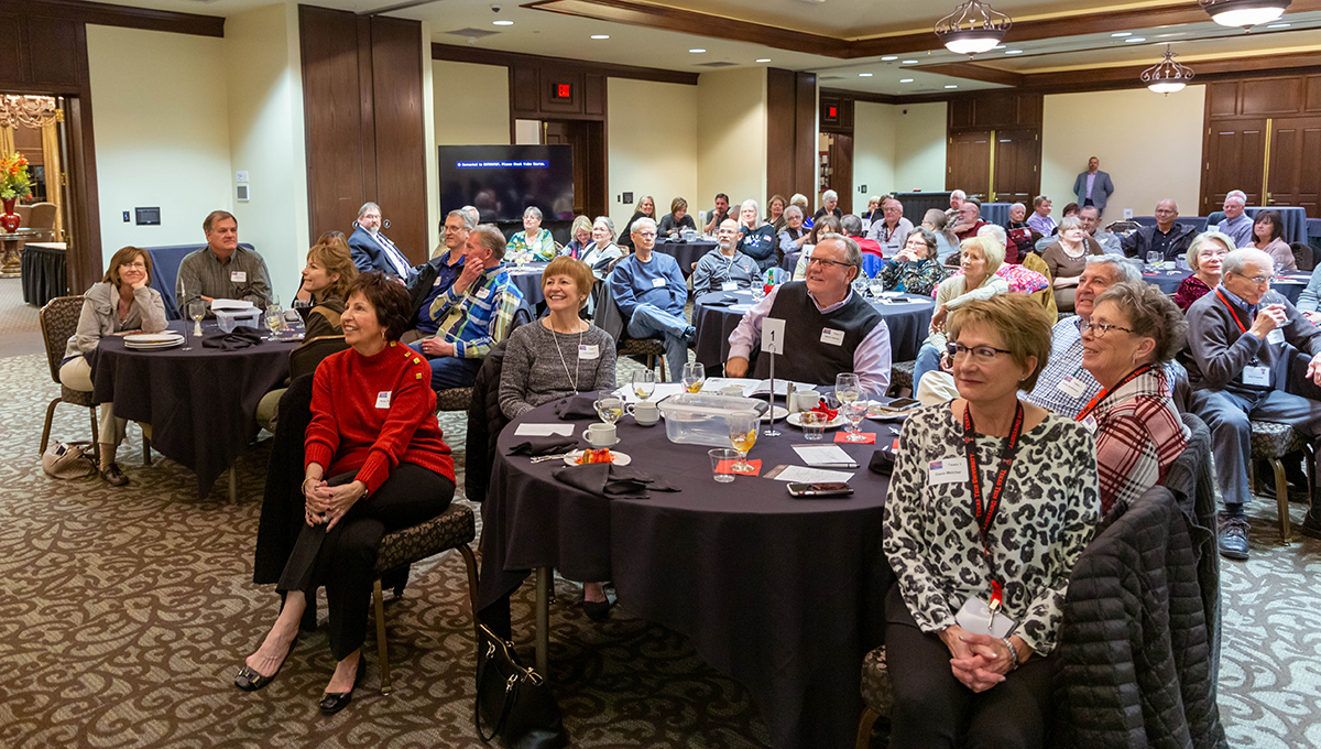 Trivia Night Emcee Jeff Klotzman A group of men and women sit around numerous round tables while looking to the front of the room